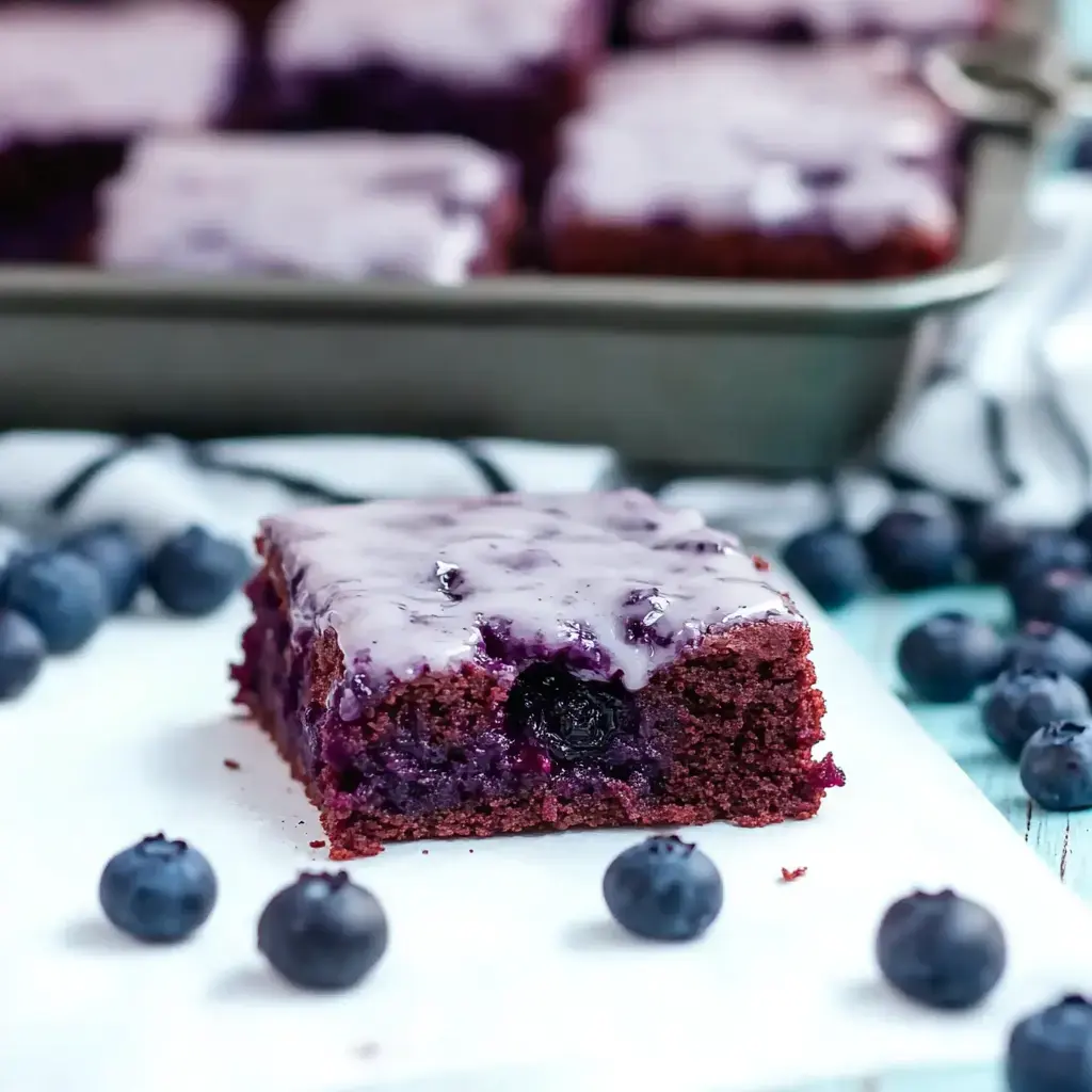 A rich, purple-frosted cake slice is displayed on a white surface, surrounded by fresh blueberries.