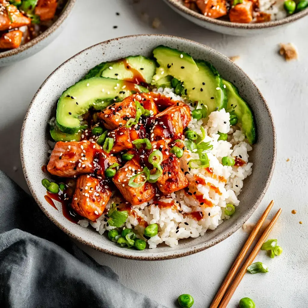 A bowl of rice topped with diced chicken, cucumber slices, green onions, sesame seeds, and drizzled with sauce.