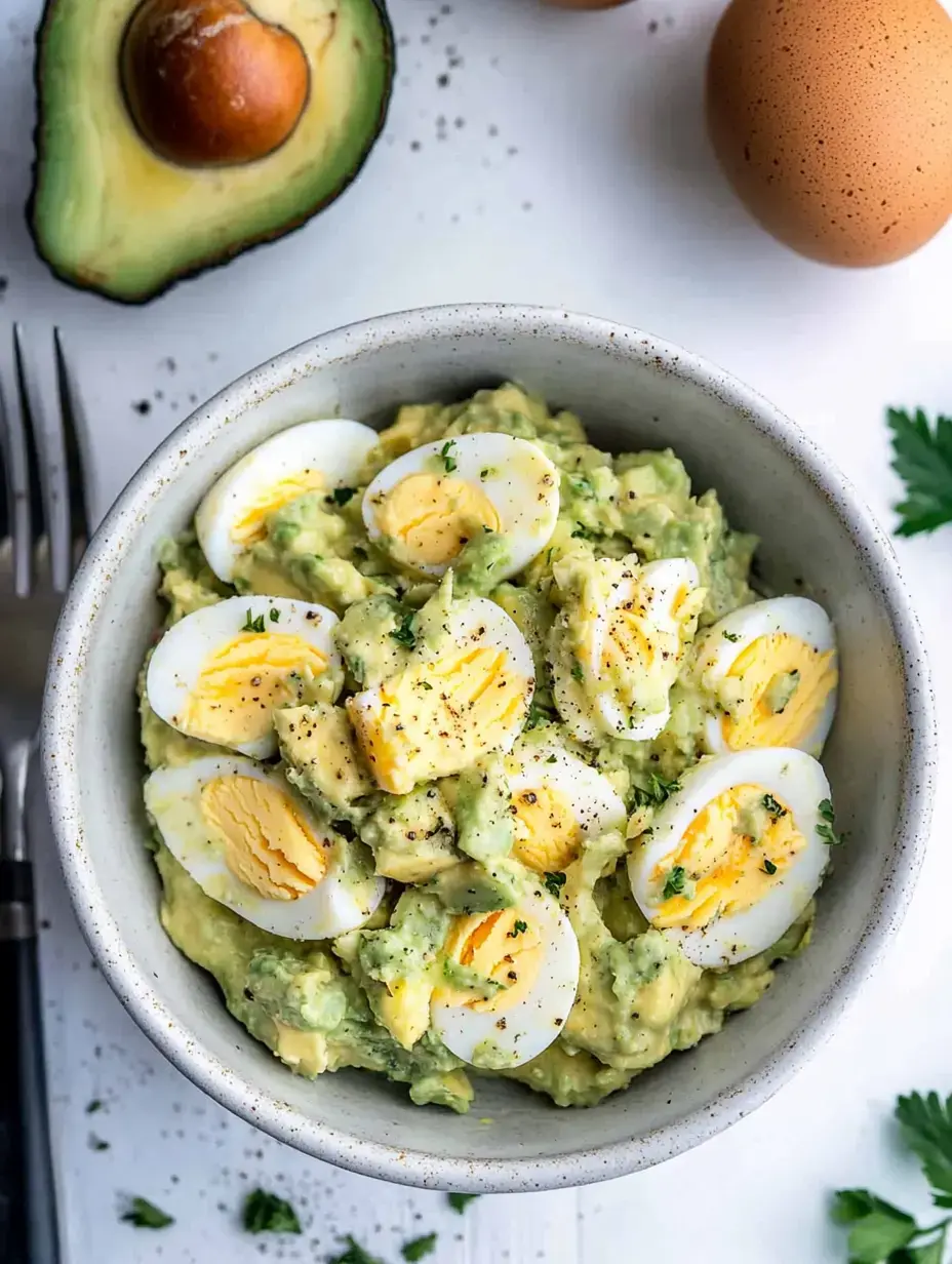 A bowl of mashed avocado mixed with chopped hard-boiled eggs, garnished with herbs and pepper, accompanied by an avocado and eggs in the background.