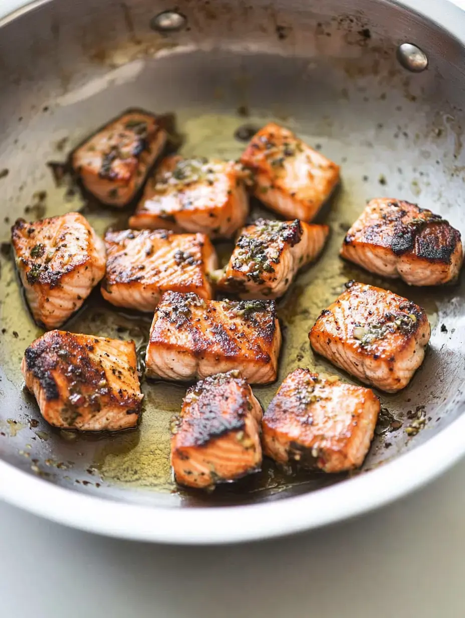 Cubes of seared salmon are arranged in a stainless-steel skillet, garnished with herbs and browned to perfection.